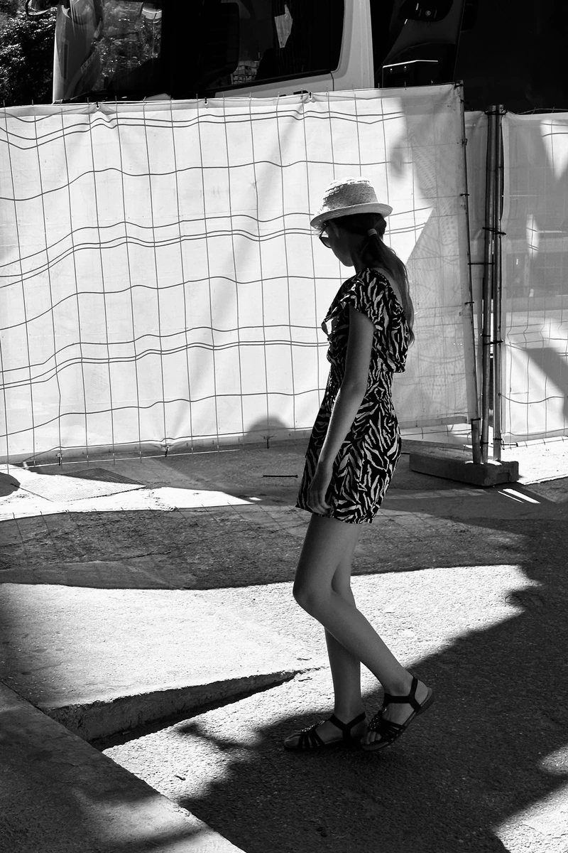 A woman in a patterned dress walks beside a sunlit construction fence