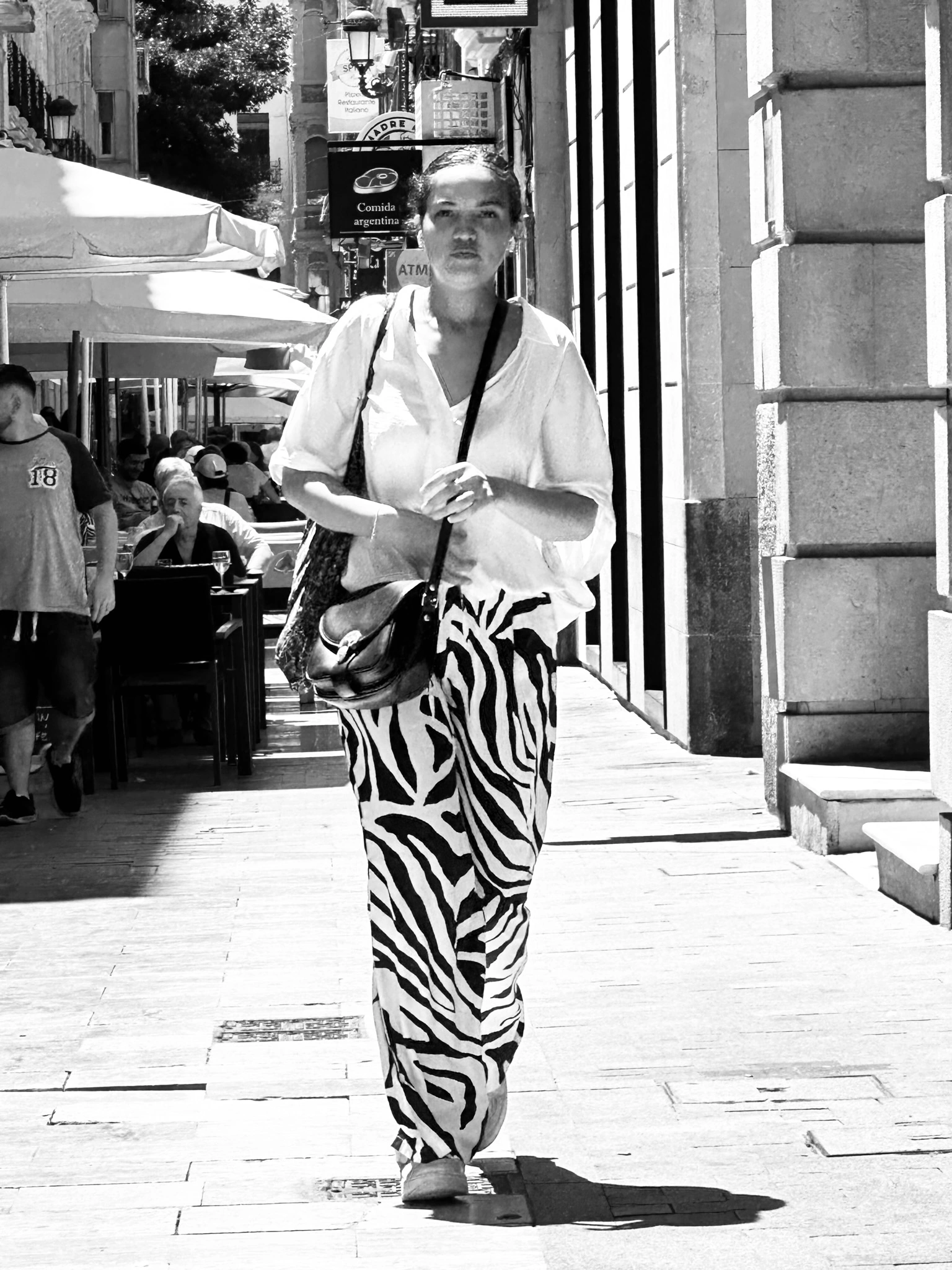A woman wearing striped trousers walks along a sunlit street