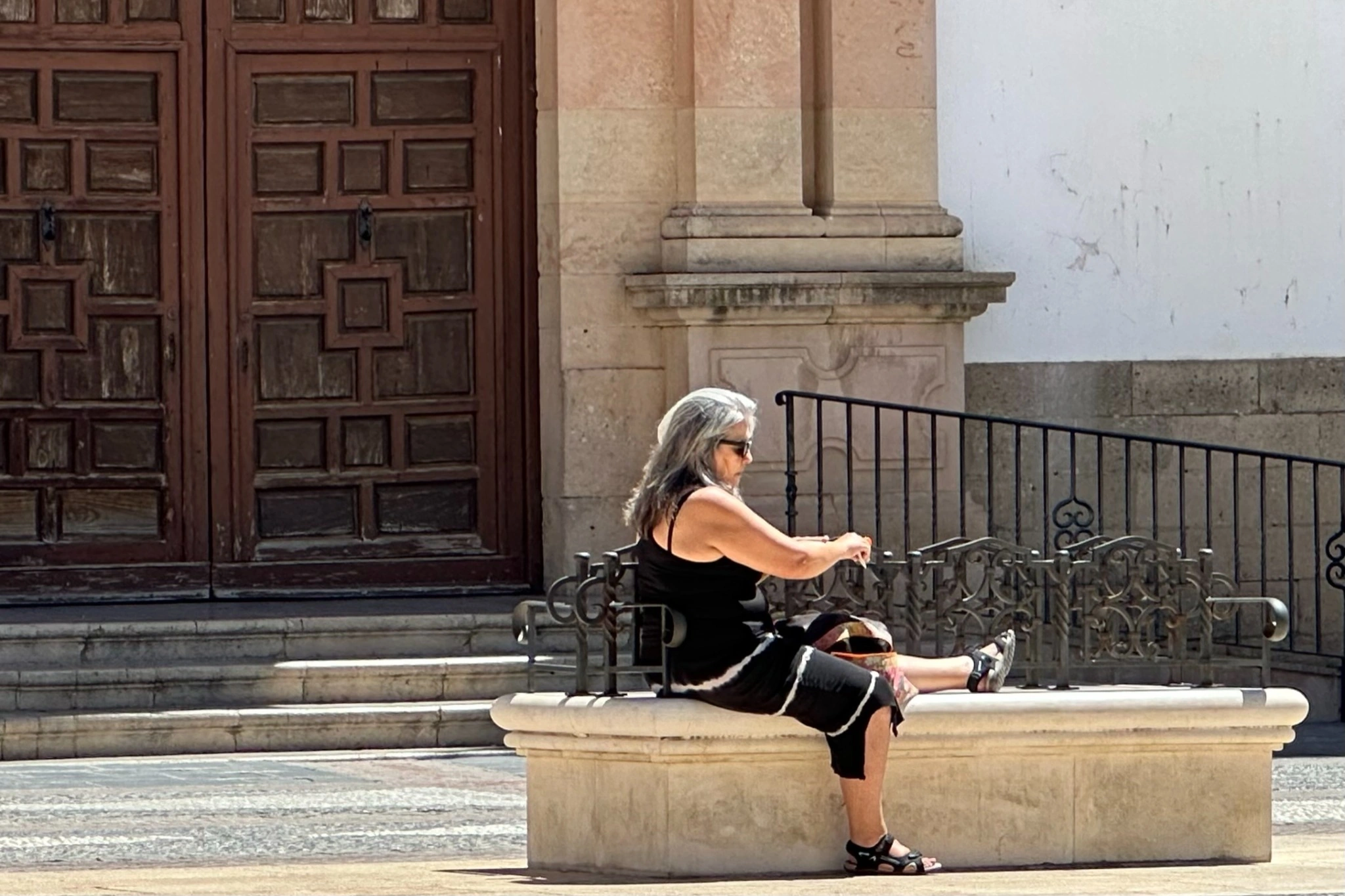 A woman seated on a stone bench, framed by a wooden doorway and iron railings