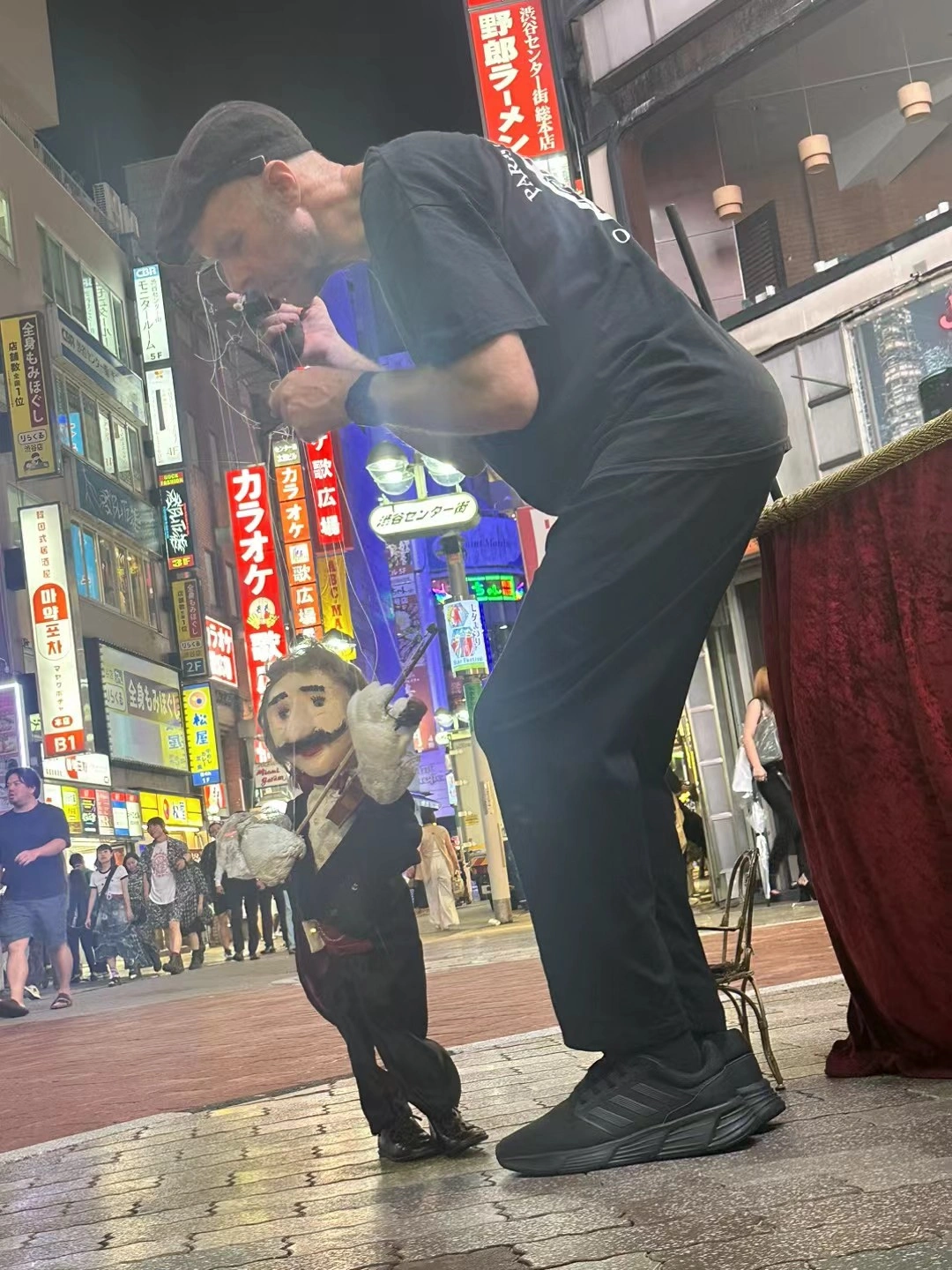 A street puppeteer leans over a marionette beneath the illuminated signs of Shibuya.