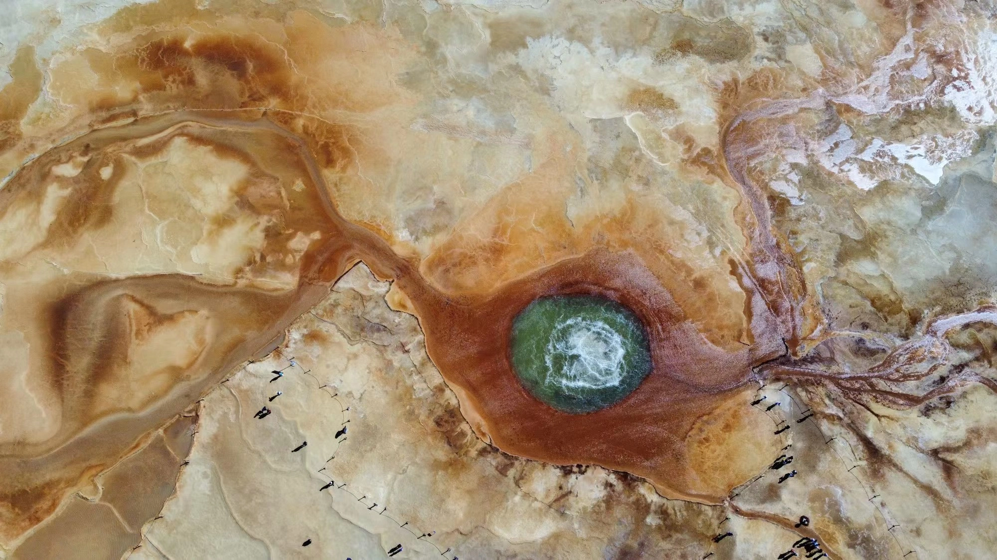 An elevated view of mineral formations surrounding a circular pool, with tiny people tracing the path.